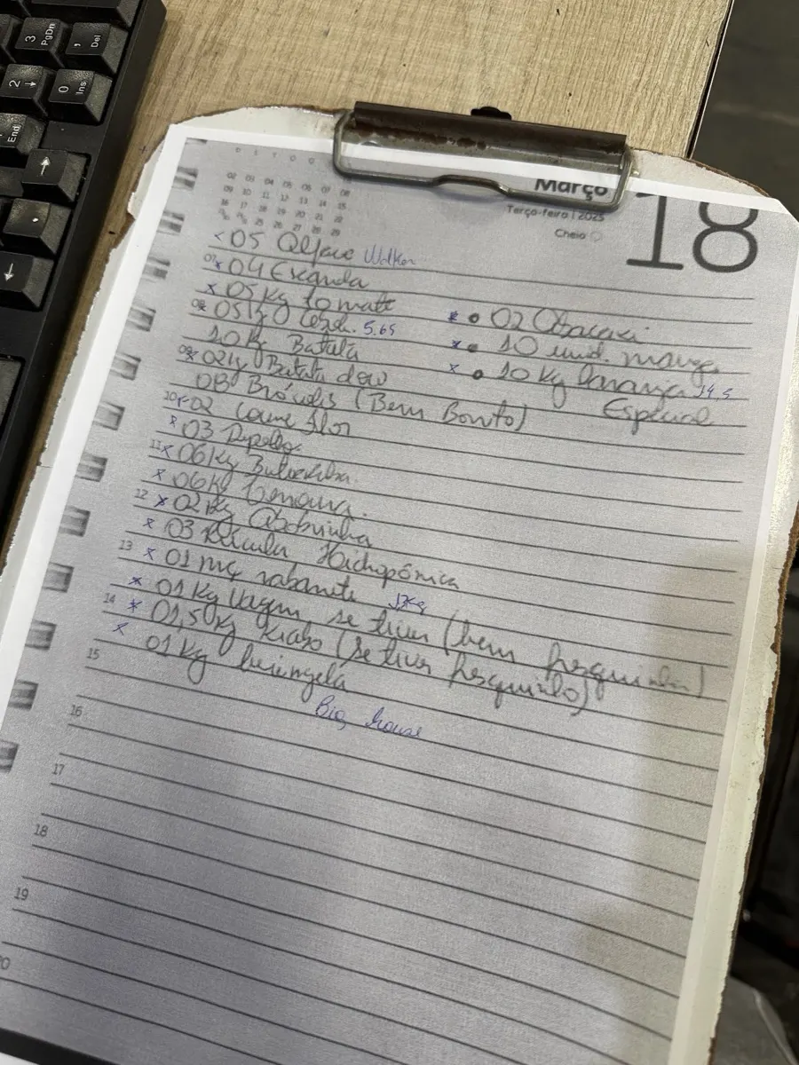 Pedido manuscrito numa folha de agenda presa a uma prancheta, sobre a mesa ao lado de um teclado
